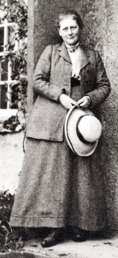 A photograph of Beatrix Potter standing outdoors in a tweed jacket and long skirt, holding a straw hat
